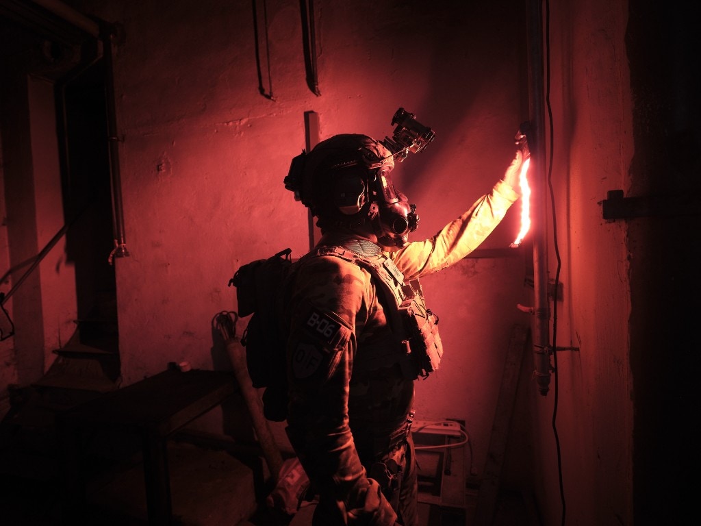 Operator in tactical gear and gas mask bending a flexible XP ALPHA 2⁺ LED strip against a pipe; deep red illumination in a dark industrial interior with NVG helmet
