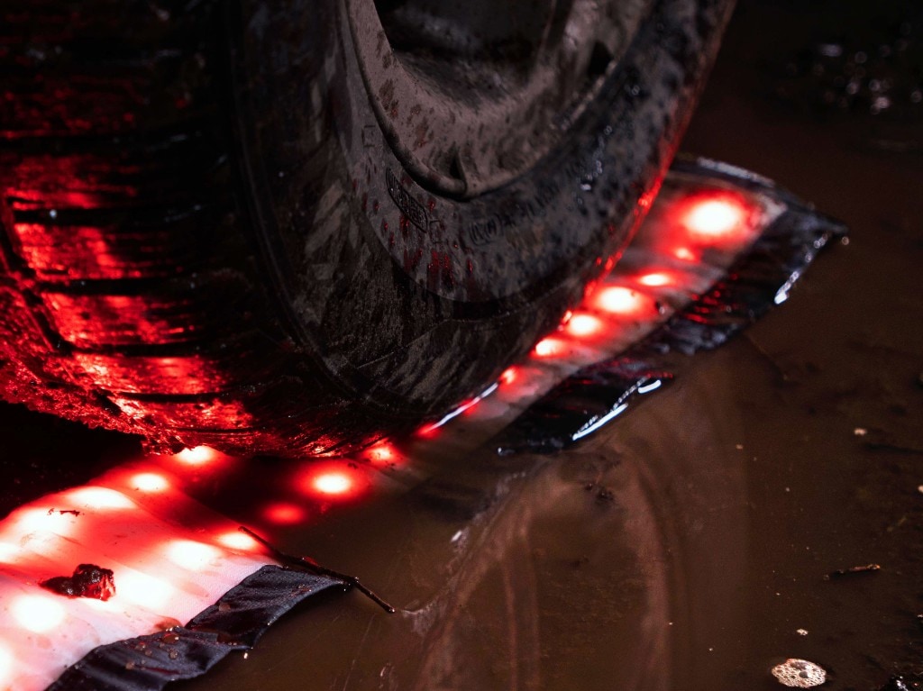 Off-road tire rolling over a flexible red LED strip on wet muddy ground at night; red glow reflecting in puddles and tread, rugged outdoor scene
