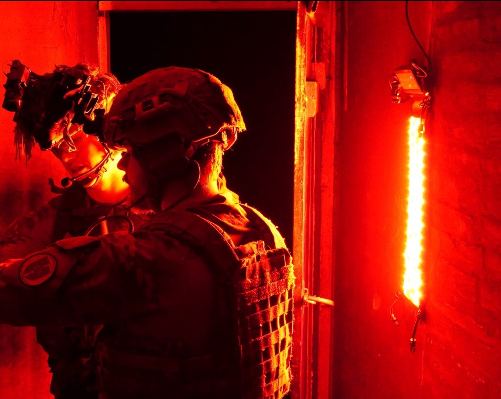 Two soldiers in tactical gear and helmets near a dark doorway; vertical red LED strip with control box on the right wall, sole light source in a blacked-out room