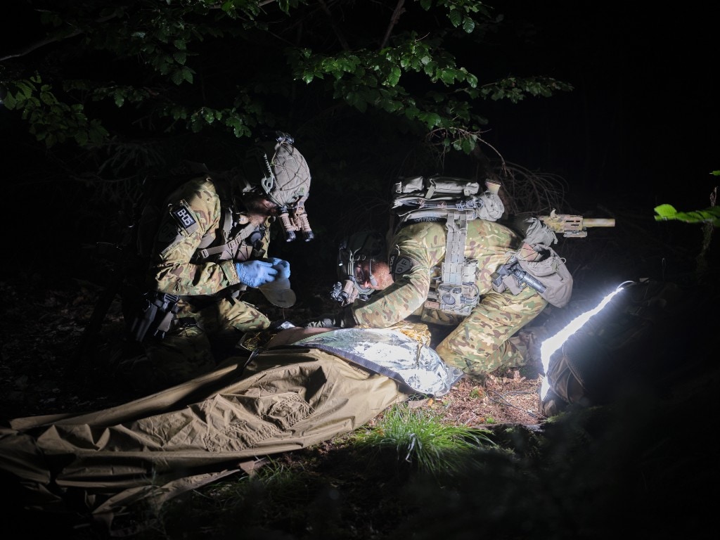 Tactical medics in multicam with NVGs treating a casualty in a bivvy bag in a forest at night; broad white portable LED panel lighting the procedure