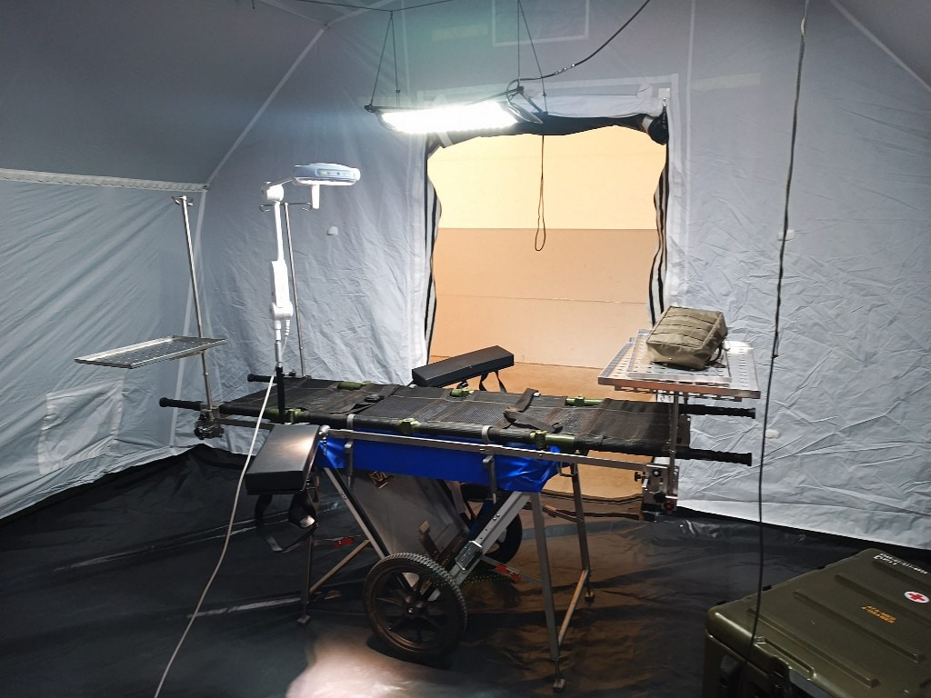 Deployable surgical gurney with side trays and IV pole inside a pale field tent; bright flat LED panel suspended from the ceiling, medical chest with red cross visible