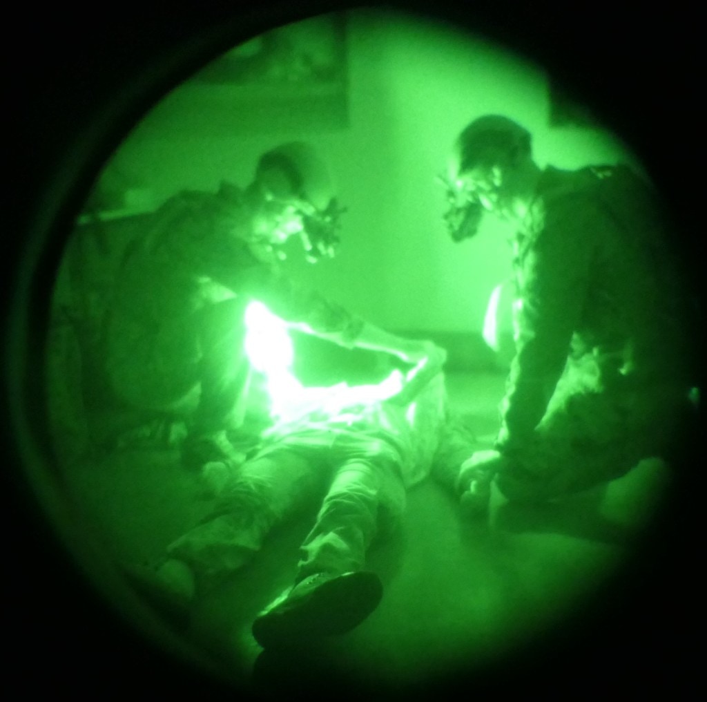 Night-vision circular green view: two helmeted tactical responders kneeling over a casualty on the floor in a dark room; intense IR bloom on the patient as seen through NVGs