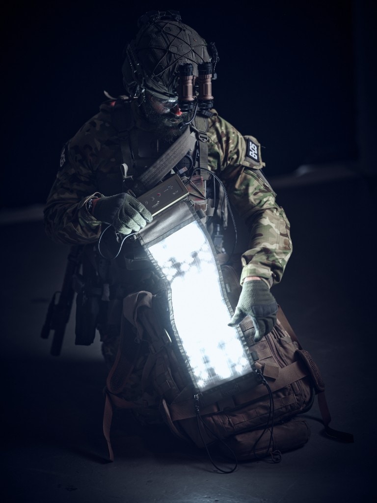 Soldier in multicam kneeling at night beside a large tan MOLLE rucksack; flexible cool-white LED panel draped over the pack with handheld DELTA-style control unit, NVGs flipped up on helmet