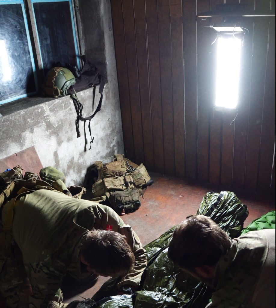 Two soldiers in multicam working over gear in a dim field room; vertical cool-white LED work light mounted on a wooden slat wall, plate carrier and helmet nearby, strong task lighting for logistics