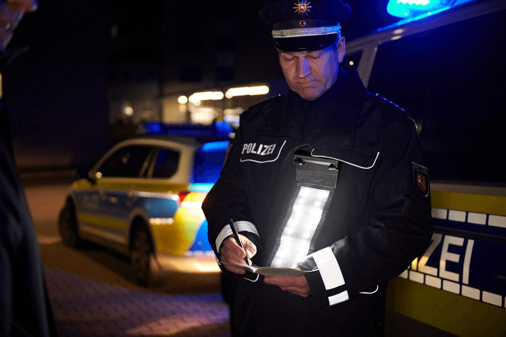 Police officer in uniform writing in a notebook at night, hands-free chest-mounted LED panel lighting the scene; patrol vehicles with blue lights in the background