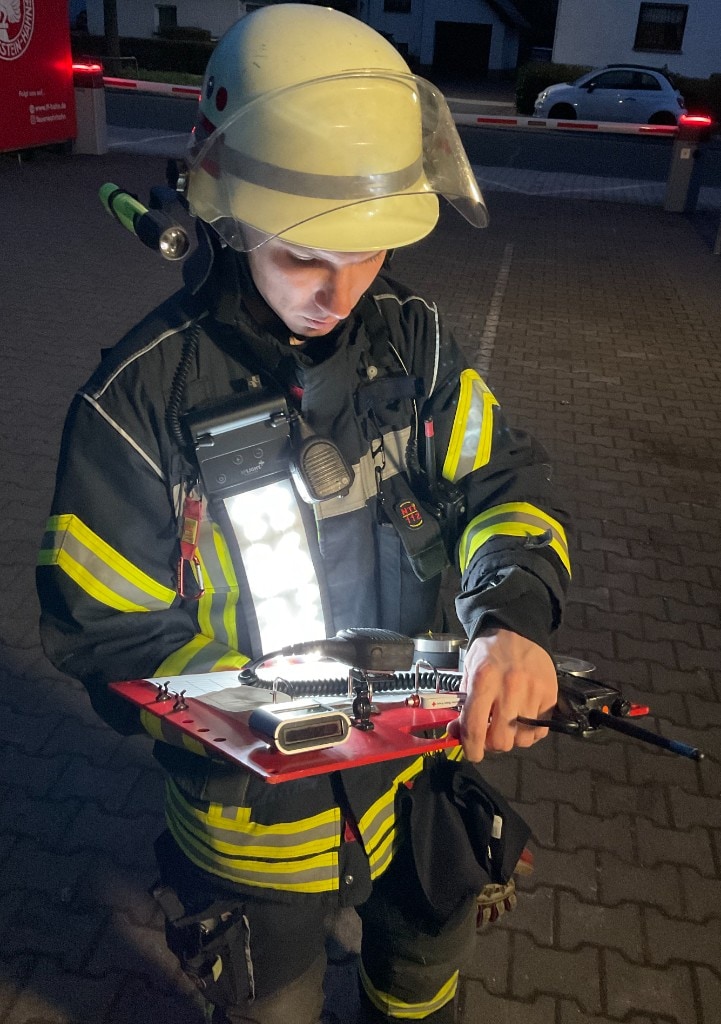 Firefighter in navy jacket with reflective stripes and white helmet at night; vertical cool-white chest-mounted LED panel lighting a red command board with radio and paperwork; cobblestones and emergency vehicle lights in background