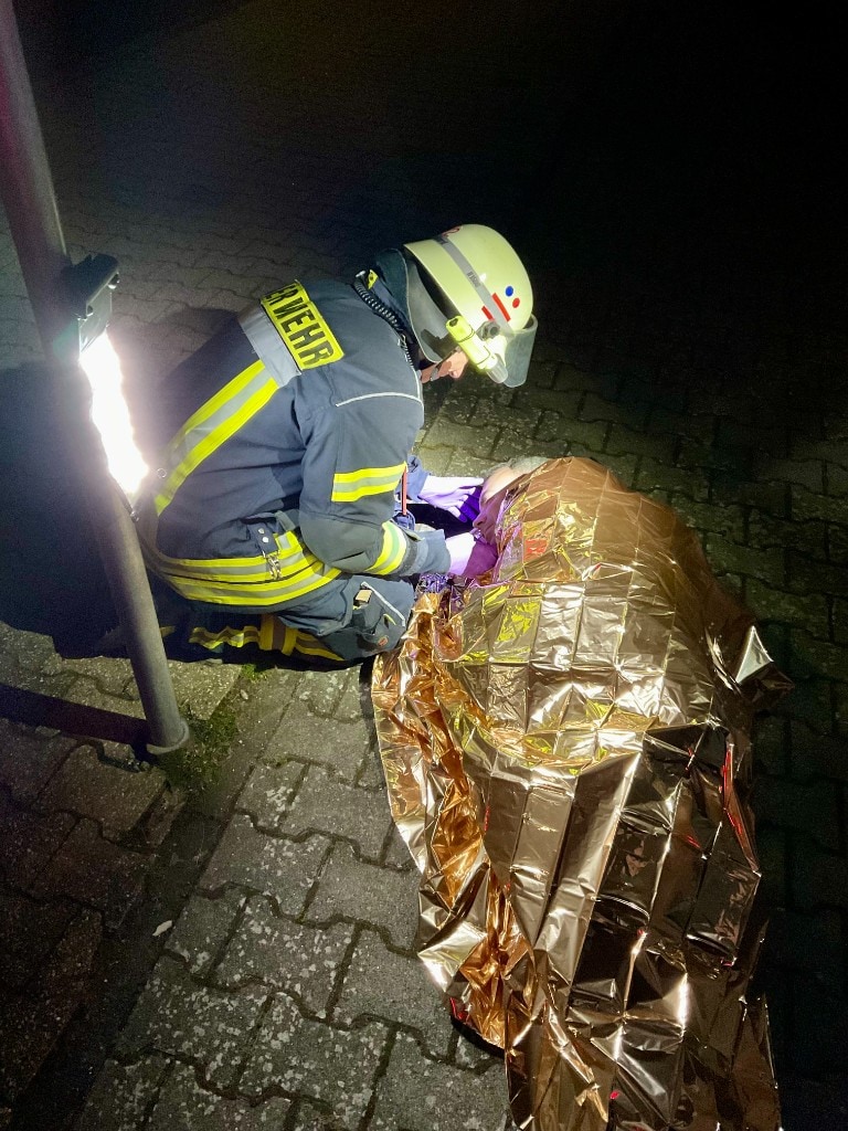 Night scene on paving stones: firefighter in FEUERWEHR jacket and white helmet kneeling over a casualty wrapped in a reflective gold thermal blanket; bright portable work light on a pole at left