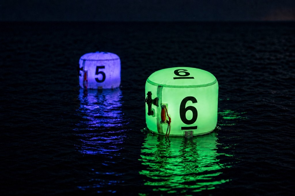 Two XP FROQ marking buoys floating on dark water at night: foreground buoy lit bright green with black number 6, background buoy lit purple with number 5, coloured reflections on the rippling surface