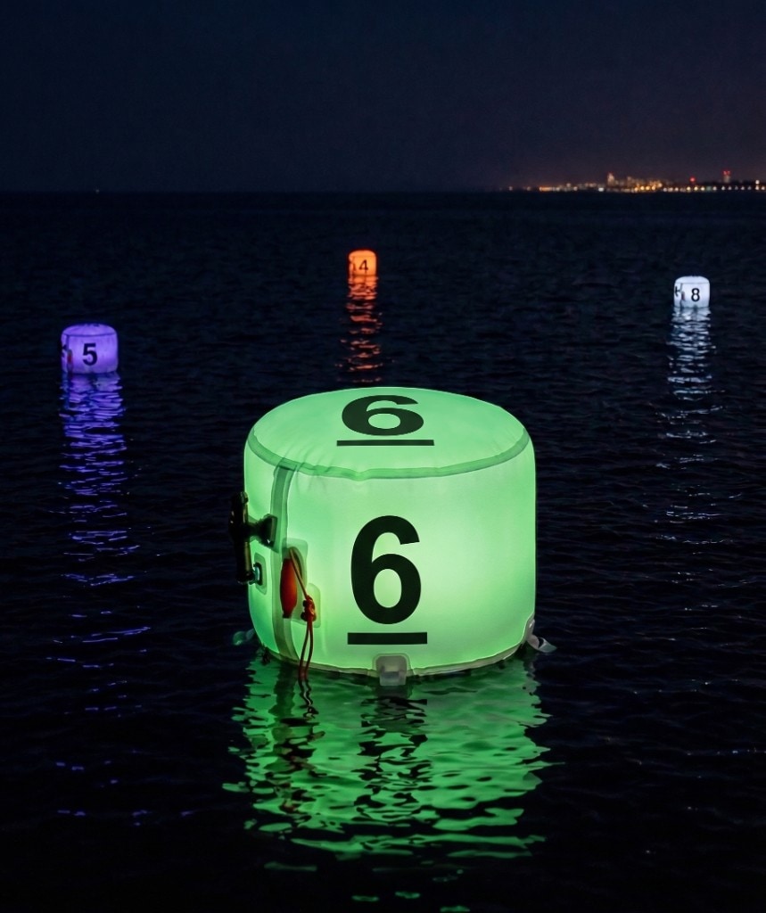 Four XP FROQ marking buoys on dark water at night in green (6), purple (5), orange (4) and white (8), spaced apart with coloured reflections; distant city lights on the horizon