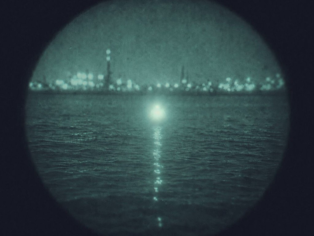 Night-vision view through circular optics on dark water: XP FROQ buoy appears as a bright IR signature on the rippling surface, with blurred city lights along the distant shoreline