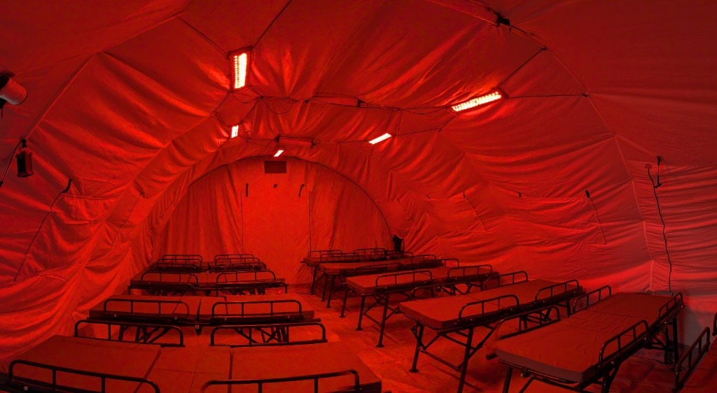 Interior of a large arched tactical tent in deep red ceiling LED light; rows of empty black-framed cots along the floor, light-colored fabric walls reflecting the red glow, low-light operations setting