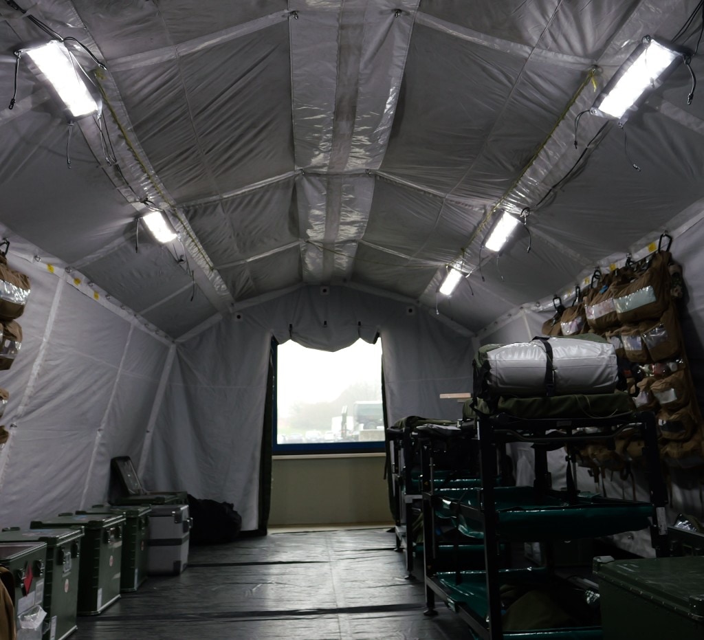 Interior of the same field tent in bright white ceiling LED lighting; central aisle toward open end, medical and operational equipment visible along the sides