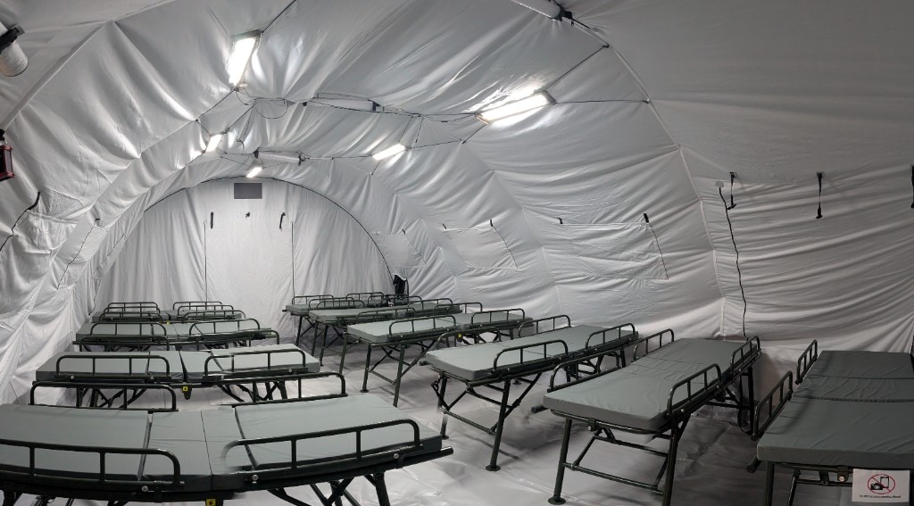 Interior of a white arched deployable shelter with integrated rectangular ceiling LED panels and routed cables; rows of olive-framed folding cots with grey mattresses and side rails, bright white stay-in-tent lighting