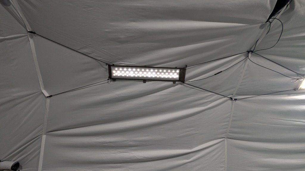 Low-angle view up at a white tent ceiling: slim horizontal rectangular LED panel with two rows of cool-white diodes, held by thin black tension lines to the fabric; power cable routed along the ceiling